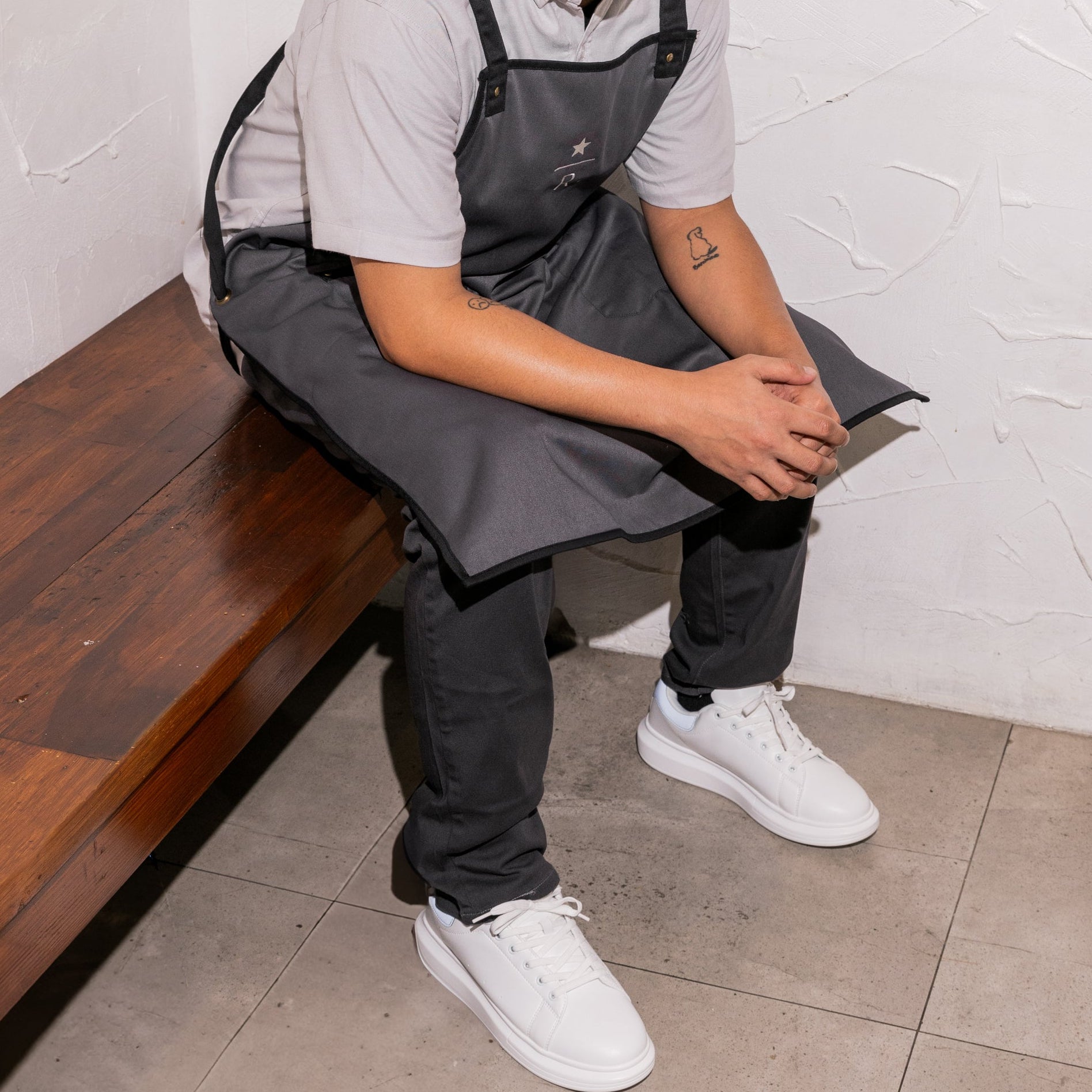 Person wearing a black apron and cap sitting on a bench against a white wall.